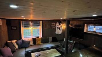 Ein gemütlicher, schummrig beleuchteter Raum mit Holzbalkendecke, einer Couch mit Kissen, einem kleinen Tisch und einem an der Wand befestigten Fernseher.