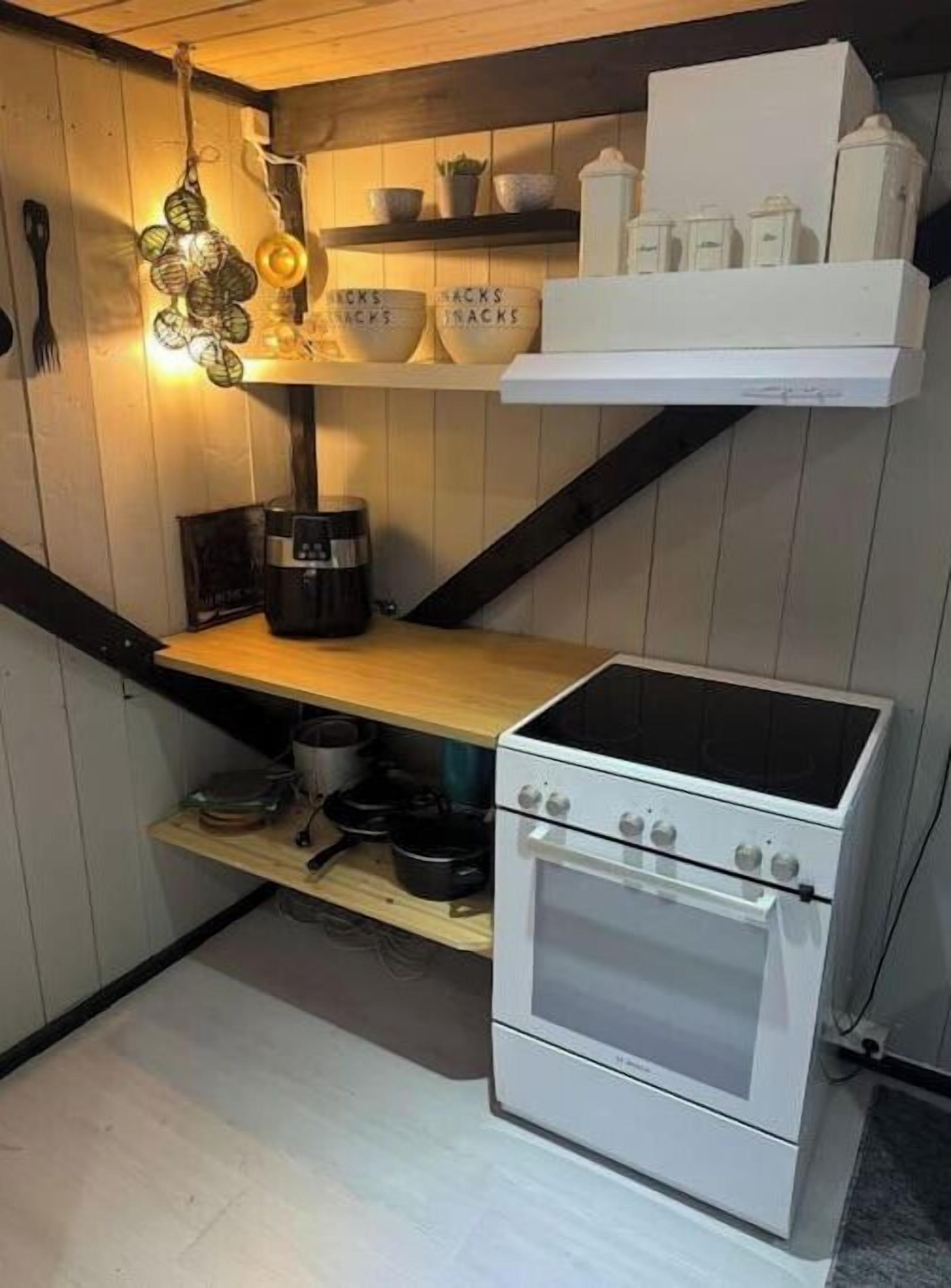 Ett kök med en spis, hyllor med köksredskap och en hängande lampa.