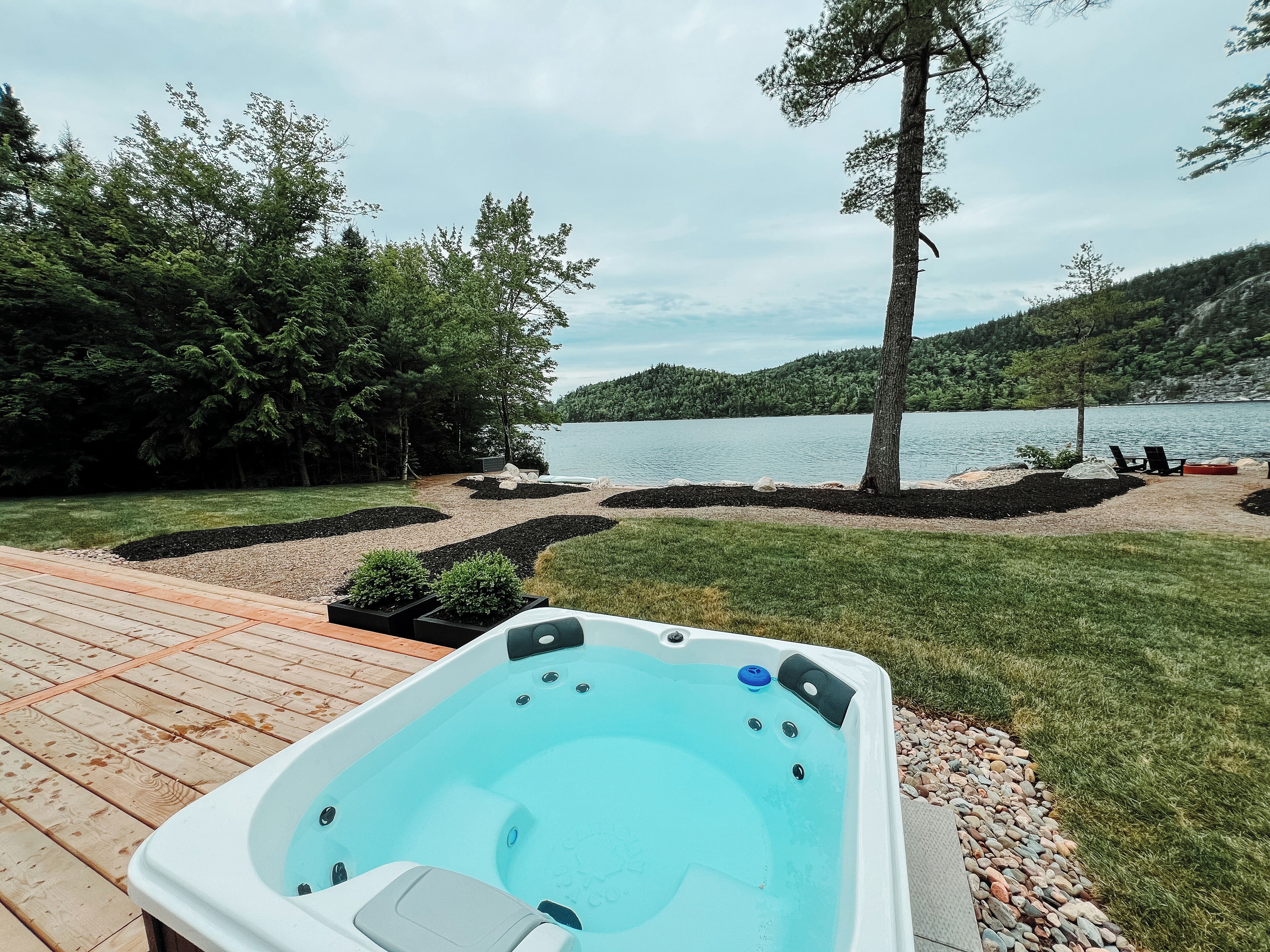 Un bain à remous sur une terrasse en bois, avec vue sur le lac, entouré d’un jardin aménagé et d’arbres.