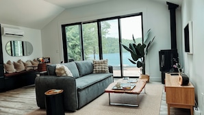 A modern living room with a grey sofa, a wooden coffee table, and a large window offering a view of a lake. - Nalu Retreat Lakeside (Porters Lake)