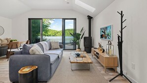 A modern living room with a large window, a sofa, a coffee table, and a television. - Nalu Retreat Lakeside (Porters Lake)