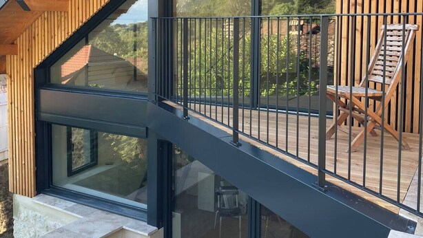 Un bâtiment moderne avec une façade en bois, de grandes fenêtres en verre et un balcon agrémenté d’une chaise.