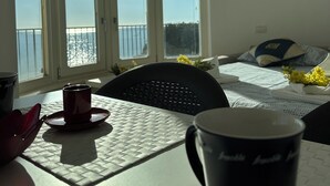 A room with a view of the sea through large windows, a bed with a blue and white pillow, and a table with a mug and a red object. - The Villino ai Tre Ponti (Sanremo)