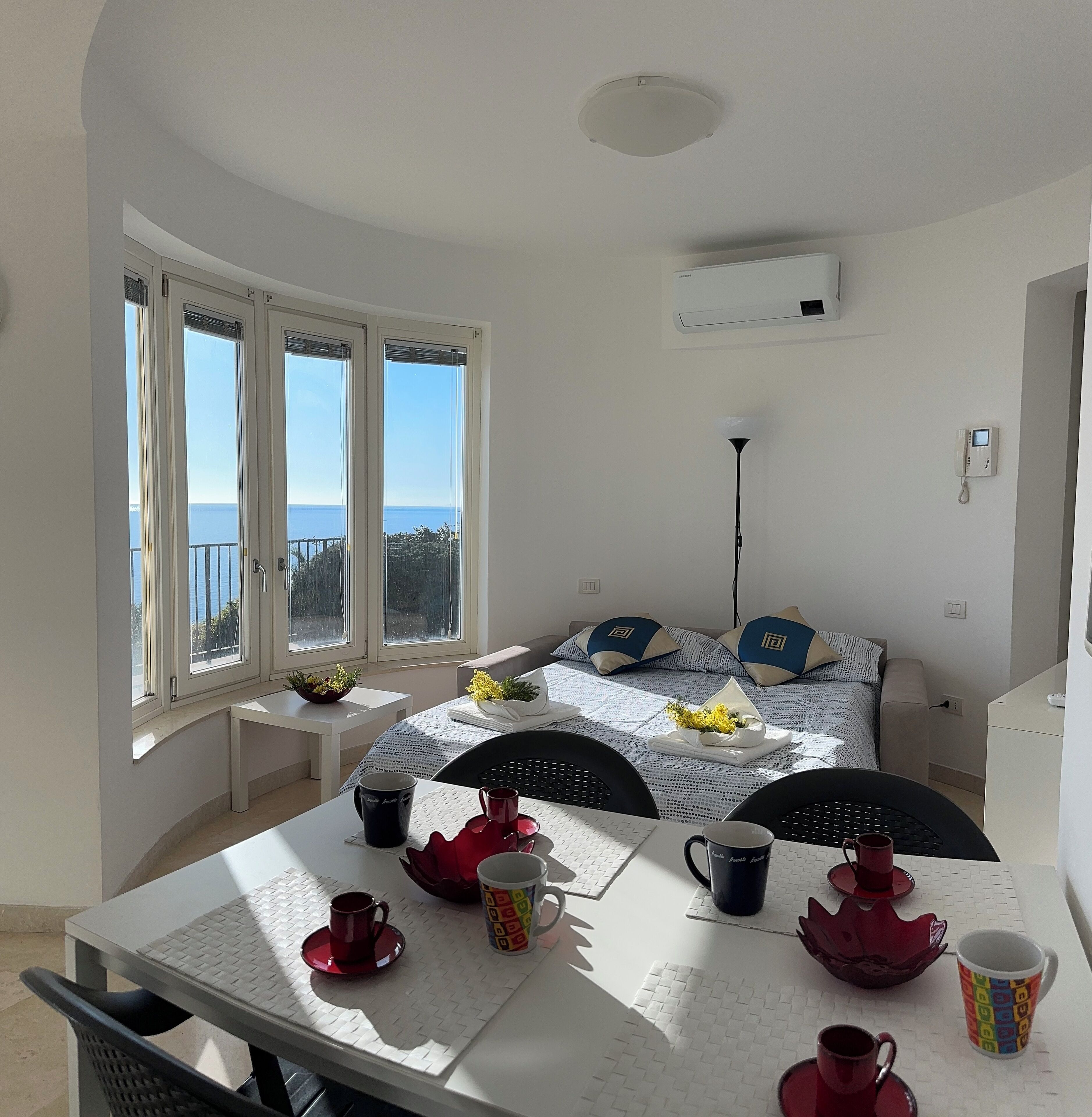 A cozy room with a bed, a table with tea cups, and a view of the sea through large windows.