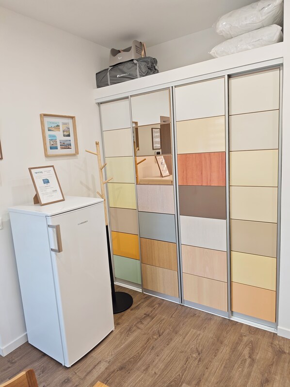 A compact room with a white refrigerator, a wooden floor, a sliding wardrobe with multiple panels, and a framed picture on the wall.