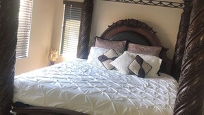 Una cama con dosel, con un cubrecolchón blanco capitonado y almohadas estampadas, un cabecero de madera y un ventilador de techo.
