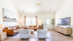 Een ruime woonkamer met een bruine leren bank, twee witte fauteuils, een houten salontafel en een houten kast.