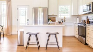 Een moderne keuken met witte kasten, een roestvrijstalen koelkast, een magnetron en een fornuis. Er staan twee barkrukken bij het eiland en een prullenbak.