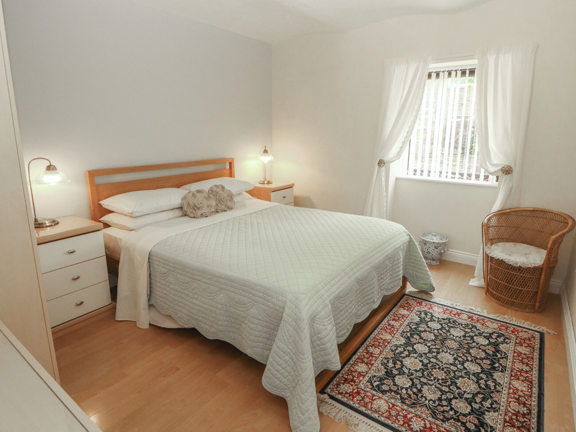 A bedroom with a bed, bedside lamps, a nightstand, a chair, a window with curtains, and a patterned rug.