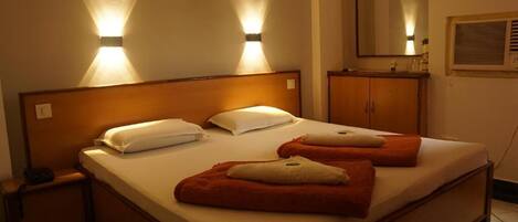 A hotel room with a large bed, two bedside lamps, a wooden headboard, a nightstand, a mirror, and a window with blinds.