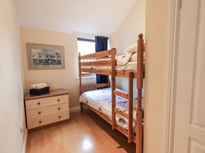 A bedroom with a bunk bed, a bedside table with a folded towel, a dresser with drawers, and a painting on the wall. - Millers Cottage (Wigton)