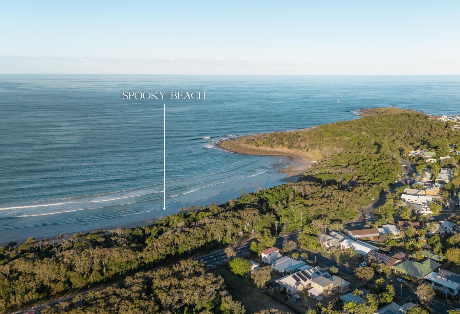 An aerial view of Spooky Beach and Paperbark, featuring a coastal landscape with a beach, dense greenery, and residential buildings.