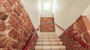 Una escalera con paredes de piedra y barandilla roja que conduce a un rellano con un ventanal pequeño.