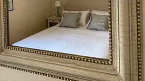 A bedroom with a large mirror, a bed with striped pillows, a nightstand with a lamp, and a ceiling light fixture. - Historic 1Bd Cottage In Heart Of Farnham (Farnham)