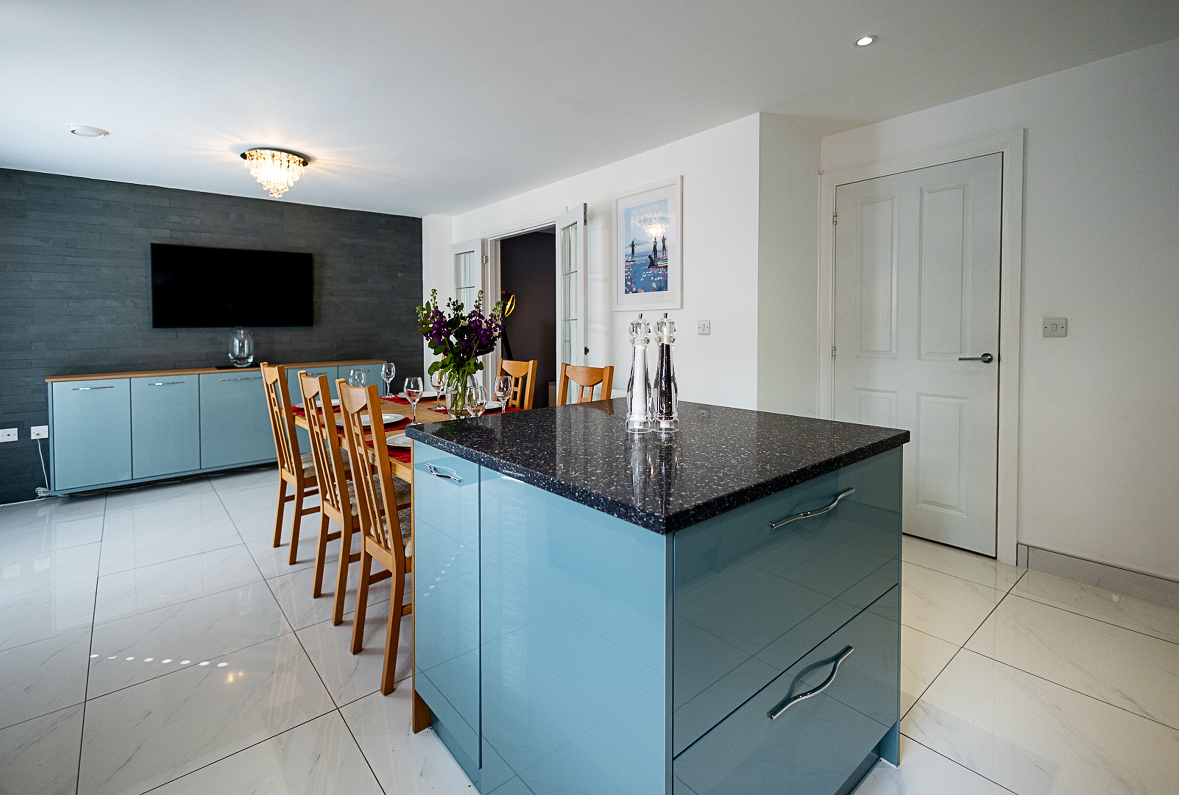 A modern kitchen with a central island, a dining area with chairs, and a flat-screen TV mounted on the wall.