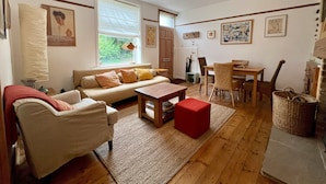 Een gezellige woonkamer met een bank, een rode poef, een houten salontafel, en een eethoek met stoelen en een tafel.