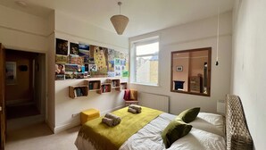 Een slaapkamer met een bed, een raam, een radiator en een muur versierd met foto’s en boeken.