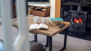 A cozy room with a wooden table, two white mugs, an open book, a remote control, and a box of tissues. A fireplace with a visible fire is in the background. - 2Bd House With Stunning Views Of The Sea And Gower (Killay)