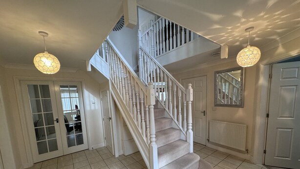 Un escalier avec des balustrades blanches et des marches recouvertes d’une moquette beige, menant à l’étage supérieur. Deux suspensions décoratives, ornées de motifs, sont suspendues au plafond.