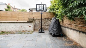 Canasta de baloncesto en un patio trasero con una pared de ladrillo, una cerca de madera y un objeto cubierto sobre ruedas.
