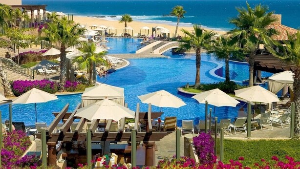 Une piscine d’un complexe hôtelier offrant une vue sur la plage et l’océan, entourée de palmiers et de chaises longues.
