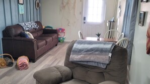 Ein Wohnzimmer mit braunem Sofa, einem beigen Sessel, einem Esstisch mit Stühlen und einer Tür mit Vorhängen.