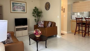 A living room with a television, a sofa, a coffee table with a floral arrangement, a ceiling fan, a clock, and a kitchen area with a microwave and chairs.