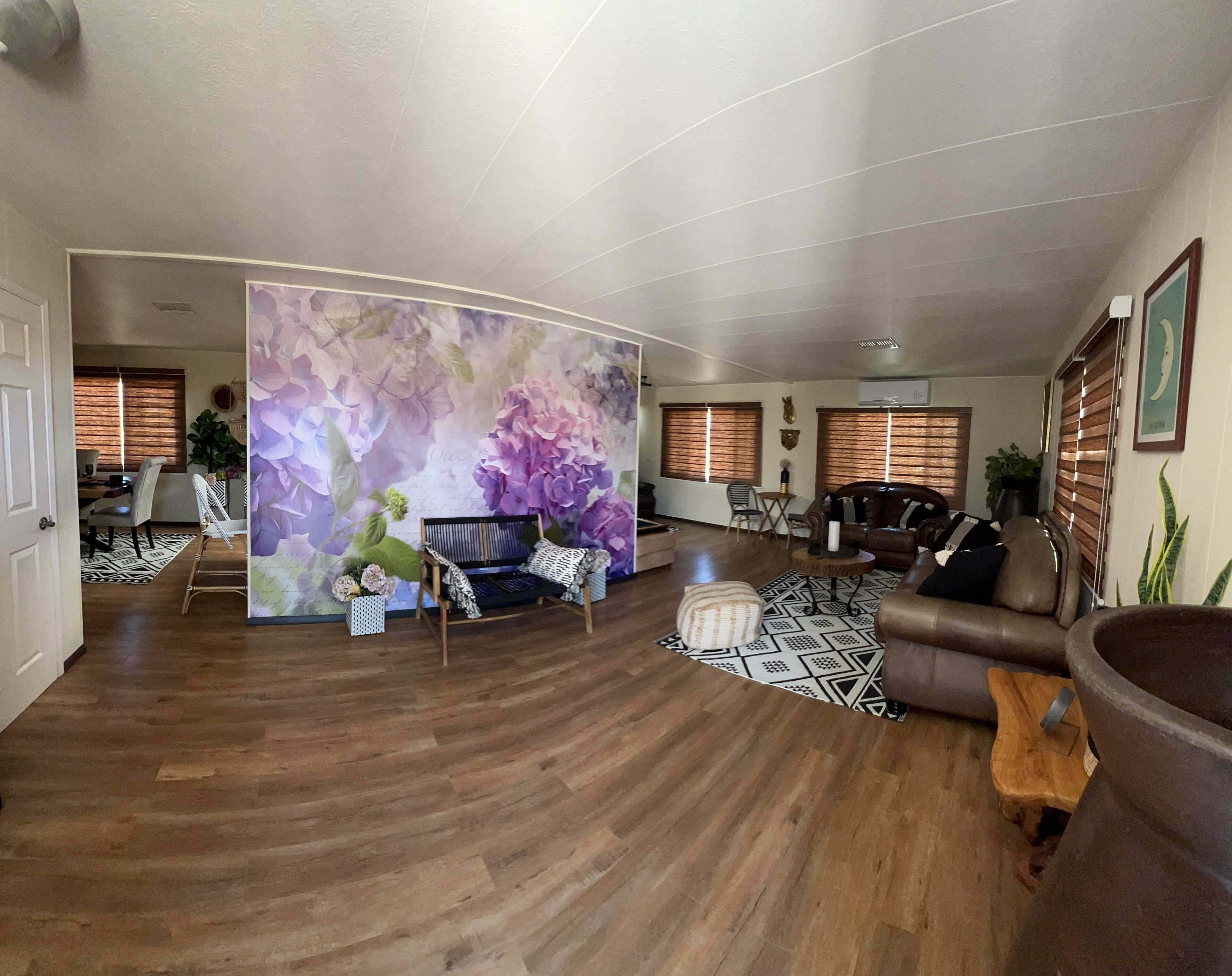 A spacious living room with a curved partition, wooden flooring, a black sofa, a patterned rug, and a large floral mural.