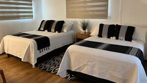 Habitación de hotel con dos camas, cada una con colcha blanca y negra, una mesita de noche con lámpara y una ventana con persianas.