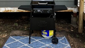 Un barbecue à gaz avec une bouteille de propane devant une maison, avec une fenêtre et une applique lumineuse fixée au mur.