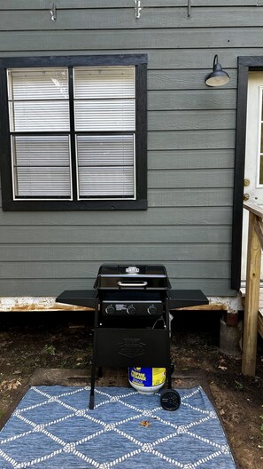 A barbecue grill with propane tank in front of a house with a window and a wall-mounted light.