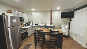 A compact kitchen with a refrigerator, stove, and microwave. There is a dining table with four chairs. A flat-screen TV is mounted on the wall.