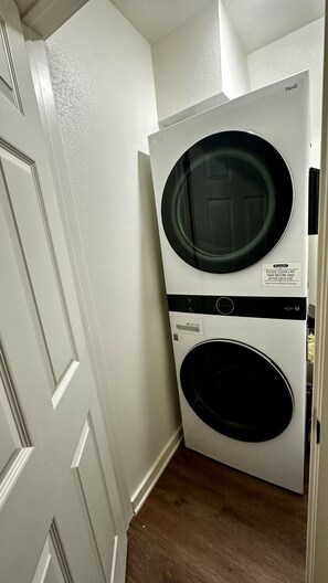 A compact washer and dryer unit with a front-loading design, placed in a corner of a room with a wooden floor and white walls.