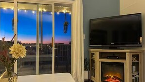 A living room with a flat-screen TV on a wooden stand, a fireplace, and a large sliding glass door leading to a balcony with string lights. - Spacious Family-Friendly Condo Near Bethany Beach (Ocean View)