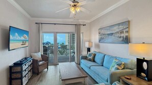 Ein geräumiges Wohnzimmer mit einer großen Couch, einem Couchtisch und einem Fernseher, der eine Strandansicht zeigt. An den Wänden sind ein Deckenventilator und ein Gemälde zu sehen.