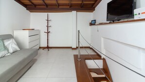 Una habitación moderna con techo de madera, un televisor de pantalla plana montado en la pared, un sofá blanco con un cojín, un perchero y una escalera con barandilla de madera.