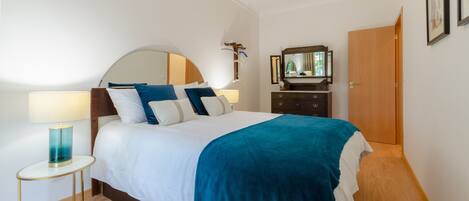 A neatly arranged bedroom with a bed, bedside table, lamp, dresser, mirror, and door.