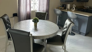 A dining area with a round table and four chairs, a kitchen with grey cabinets and a microwave, and a window with curtains. - Sea breeze retreat (Falmouth)