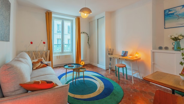 En moderne stue med sofa, sofabord og skrivebord med stol. Der er et vindue med gardiner, en hængende lampe og et billede på væggen.