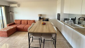 A modern kitchen with white cabinets, a wooden dining table, and a sofa. - Beautiful 2-bedroom apartment with sea view - swimming pool - tennis court (Villeneuve-Loubet)
