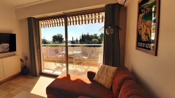 A living room with a sofa, a TV, a balcony with a table and chairs, and a poster on the wall. - Beautiful 2-bedroom apartment with sea view - swimming pool - tennis court (Villeneuve-Loubet)