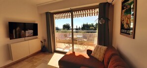Un salon avec un canapé, un téléviseur, un balcon avec une table et des chaises, et une affiche accrochée au mur.