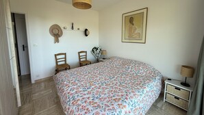 A bedroom with a large floral bedspread, two wooden chairs, a hanging lamp, a framed picture, and a small bedside table with a lamp. - Beautiful 2-bedroom apartment with sea view - swimming pool - tennis court (Villeneuve-Loubet)