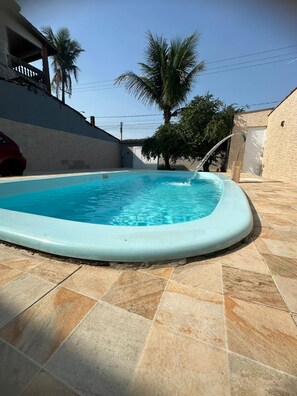 A swimming pool with a curved edge, surrounded by tiled flooring and a white wall. - Casa Paraíso em Família - Praia Grande (Praia Grande)