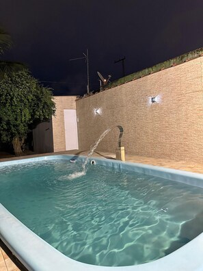 A swimming pool with a water fountain feature, surrounded by a brick wall and a tree. - Casa Paraíso em Família - Praia Grande (Praia Grande)