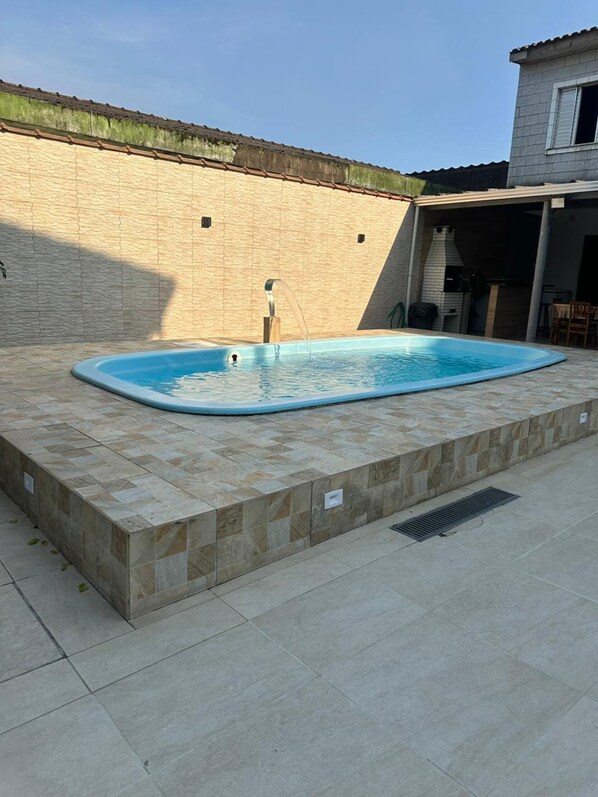 A swimming pool with a tiled deck, a brick wall, and a covered patio area. - Casa Paraíso em Família - Praia Grande (Praia Grande)