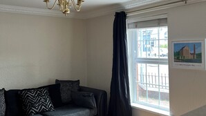 Ein Wohnzimmer mit einem dunkelgrauen Sofa, einem Holztisch, einer Kristallleuchtre und einem Fenster mit Blick auf ein Gebäude.