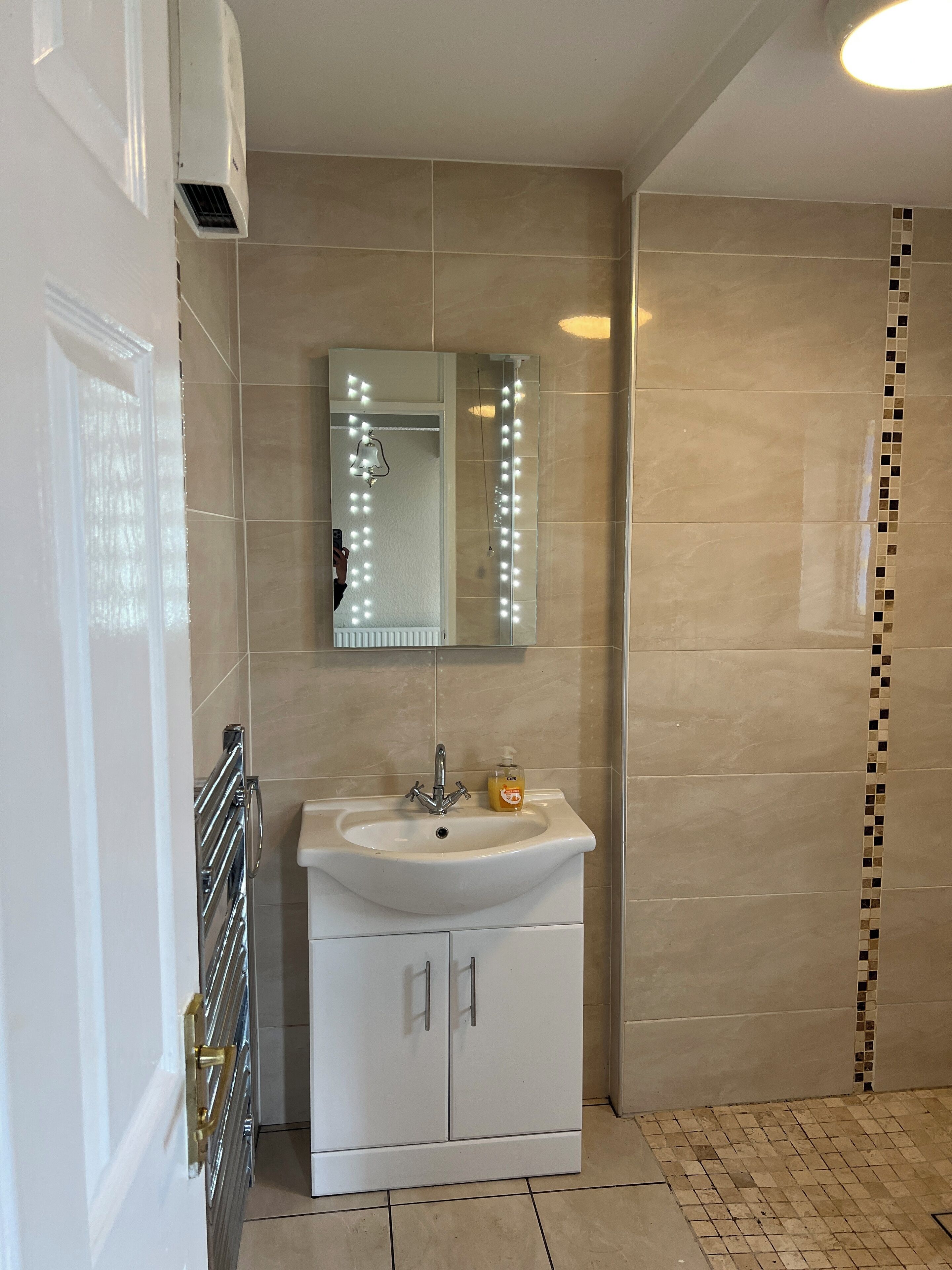 Un baño con lavamanos blanco, espejo con iluminación, ducha con pared de azulejos y piso de azulejos.