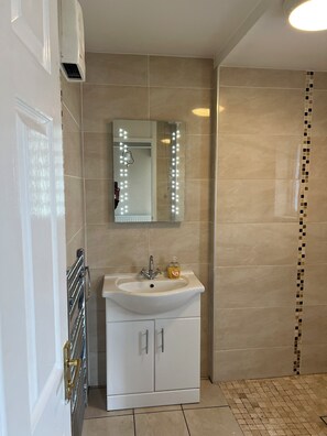 A bathroom with a white sink, a mirror with lights, a shower with a tiled wall, and a tiled floor.