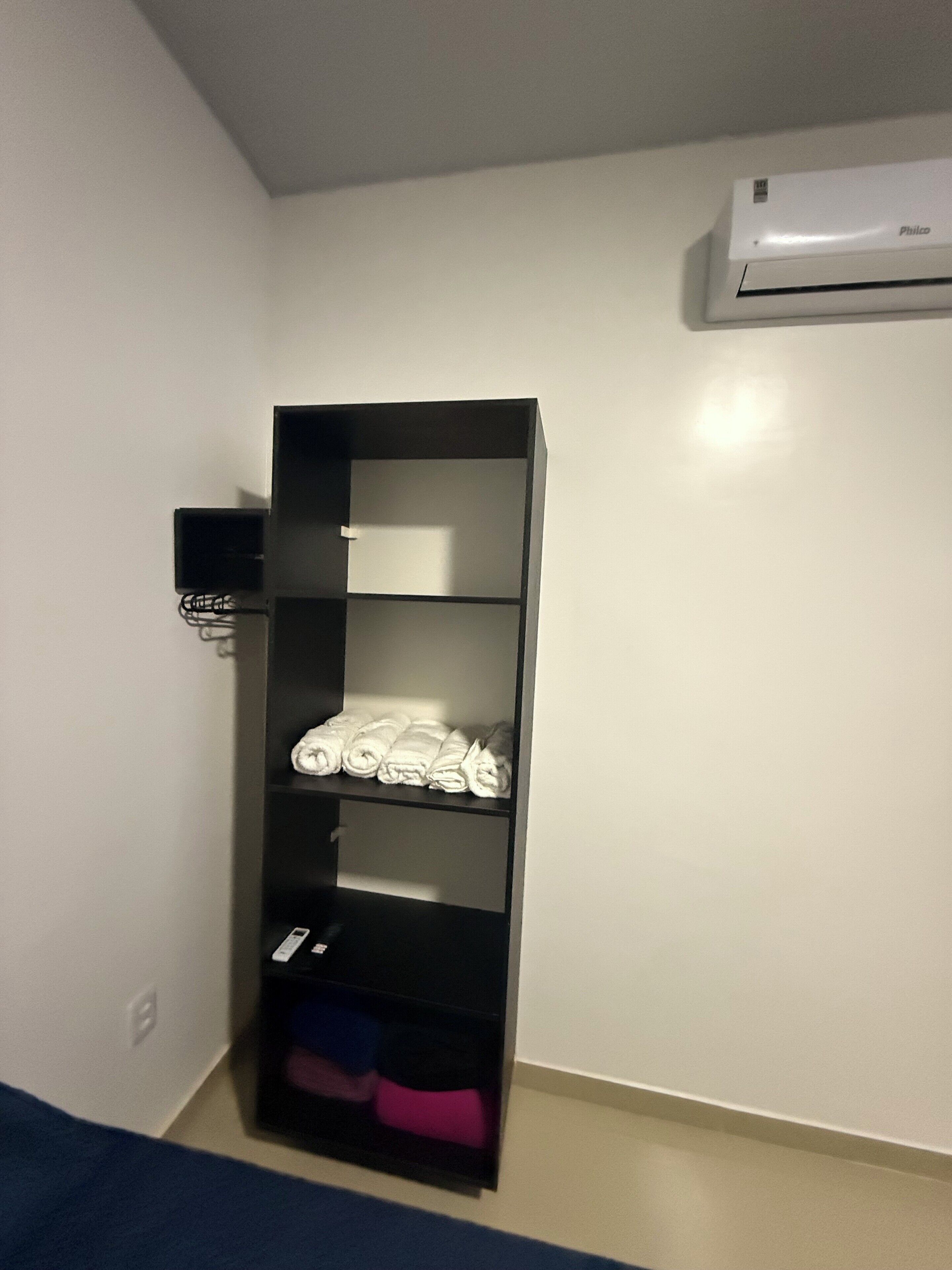 A room with a black storage unit against the wall, a white air conditioning unit above it, and a blue bed in the foreground.